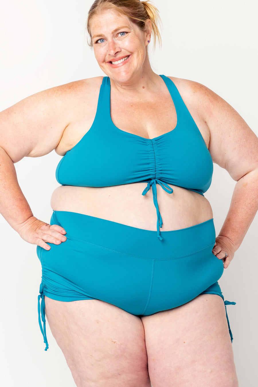 Front view of a smiling blonde model posing with hands on hips wearing high-waisted teal Superfit Hero swim booty shorts.