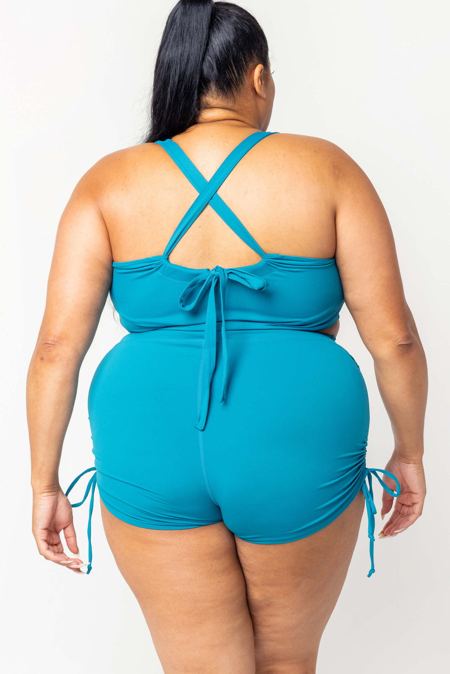 Rear full-body view of a model in teal high-waisted swim booty shorts, showcasing the smooth coverage and side drawstring ties.