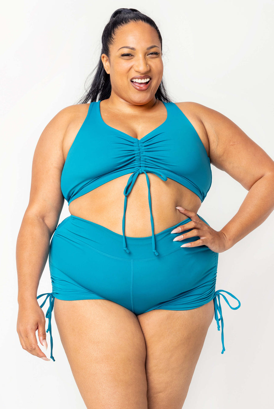 Front view of a smiling plus-size model posing with hand on hip wearing teal high-waisted swim booty shorts and a cinched bikini top.