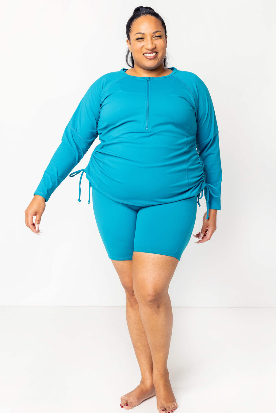 Full-body shot of a model wearing the teal long sleeve swim rash guard cinched at the sides paired with matching teal swim shorts.