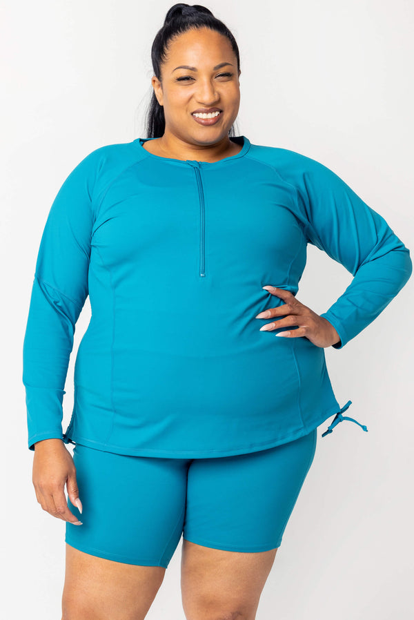 Front view close-up of a smiling plus-size model wearing the teal Superfit Hero long sleeve rash guard with half-zip collar and side drawstring ties.