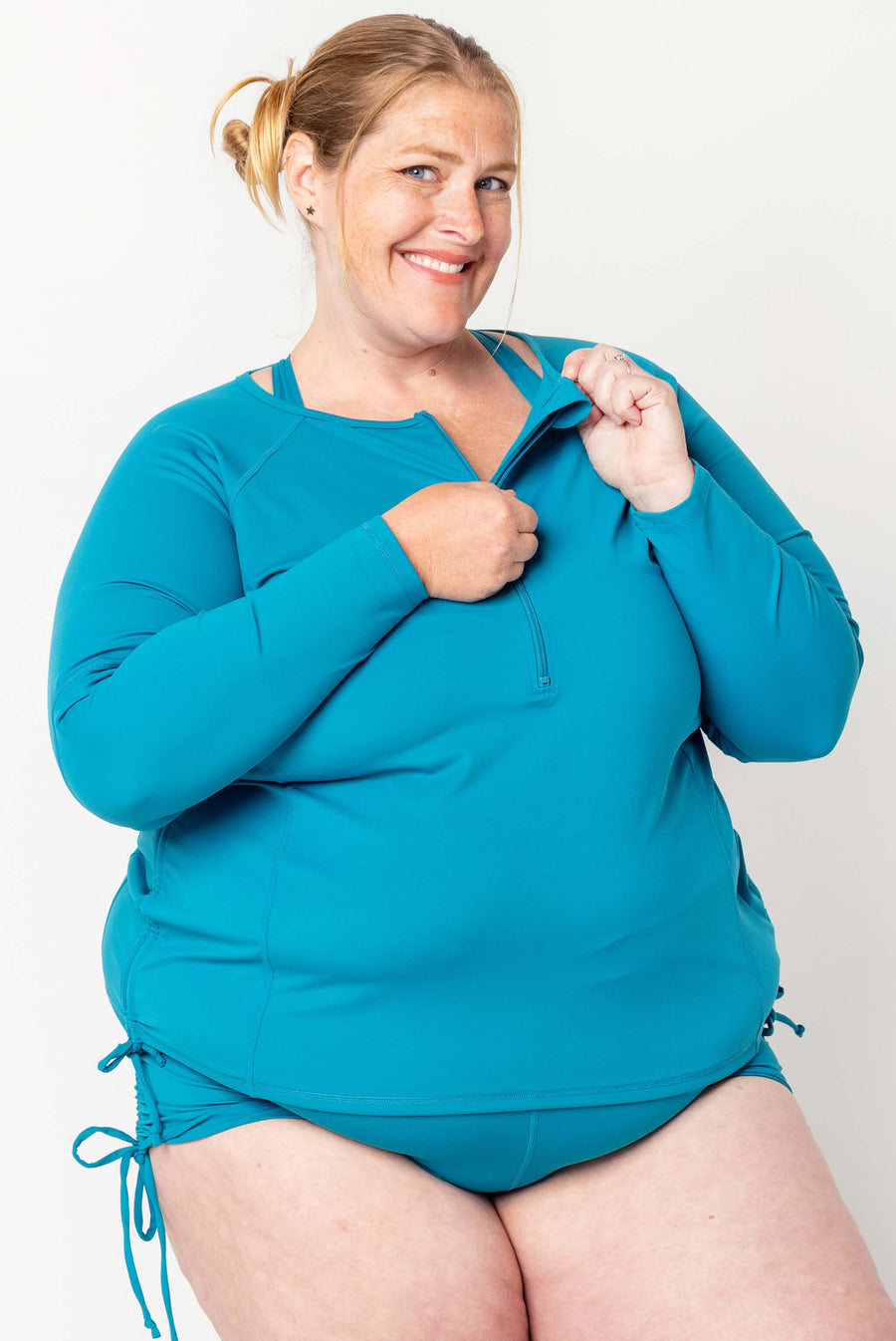 Front view of a smiling blonde model adjusting the front half-zipper on the teal long sleeve UV swim rash guard.