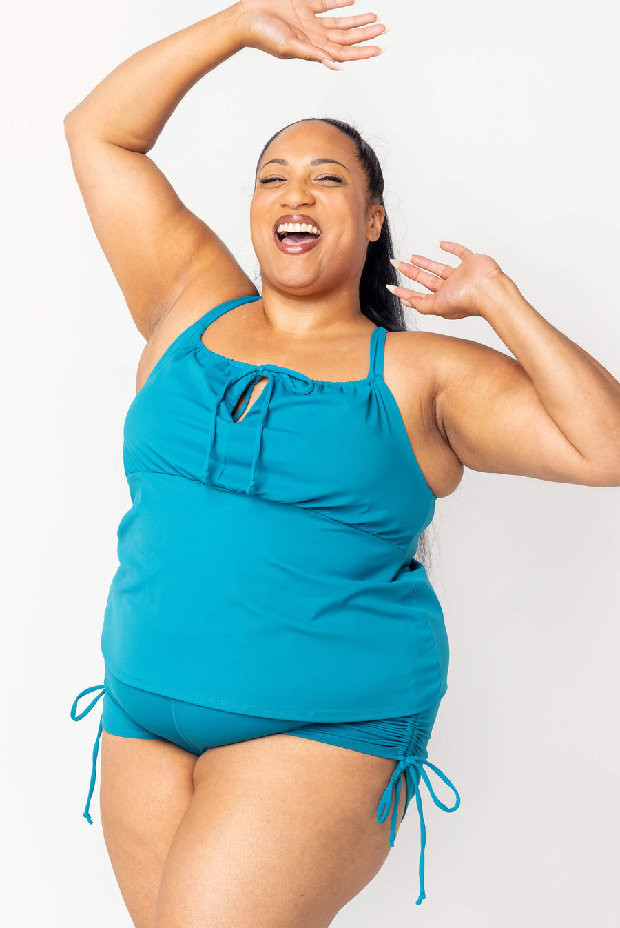 Front view of a model laughing with arms raised, showcasing the teal tankini top with front keyhole cutout paired with matching swim shorts.