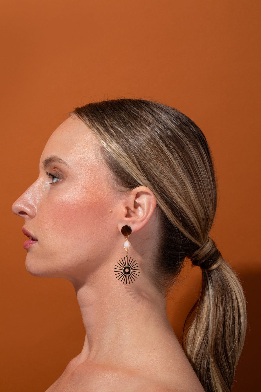 Side profile view of a model showcasing the lightweight Tribe and Temple Isela drop earring featuring a genuine pink jade stone and gold starburst charm, set against a solid warm brown studio background.