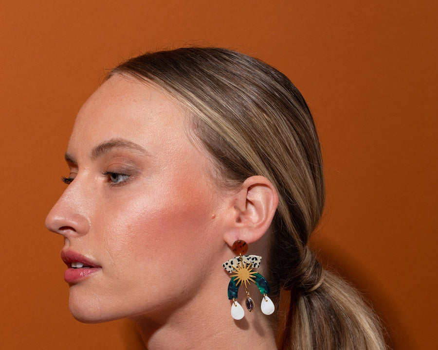 Side profile view of a model wearing a lightweight Tribe and Temple Maribel statement earring with hand-finished resin and glass drops, set against a solid warm brown studio background.