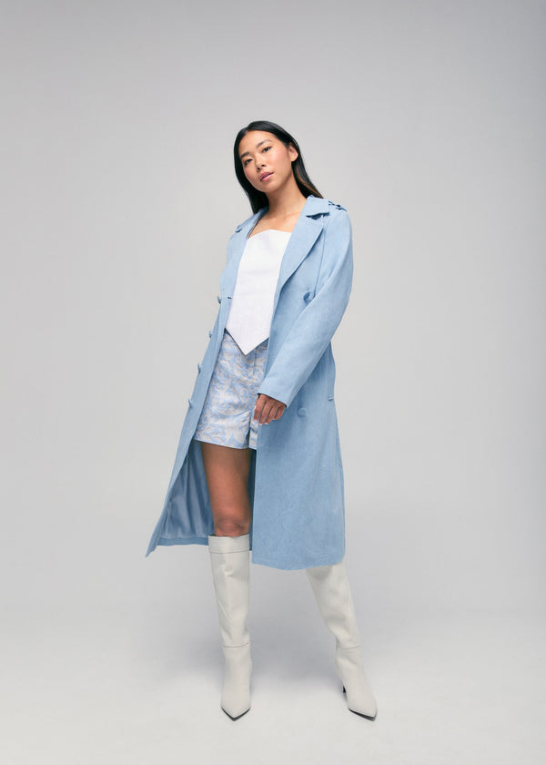 Full-length view of a model styling the light sky blue Tamika denim trench coat open over a white corset top, patterned shorts, and knee-high white boots.