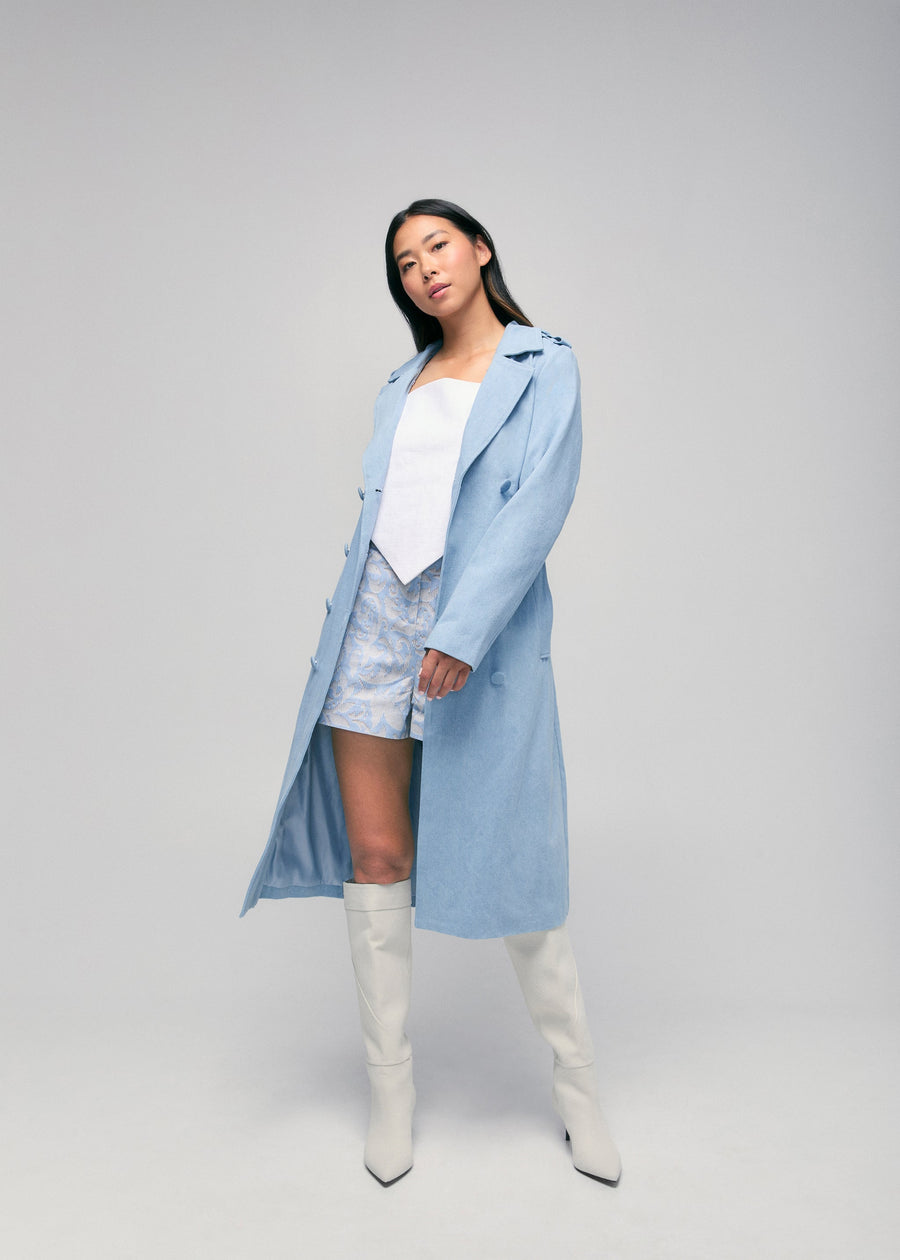 Full-length view of a model styling the light sky blue Tamika denim trench coat open over a white corset top, patterned shorts, and knee-high white boots.