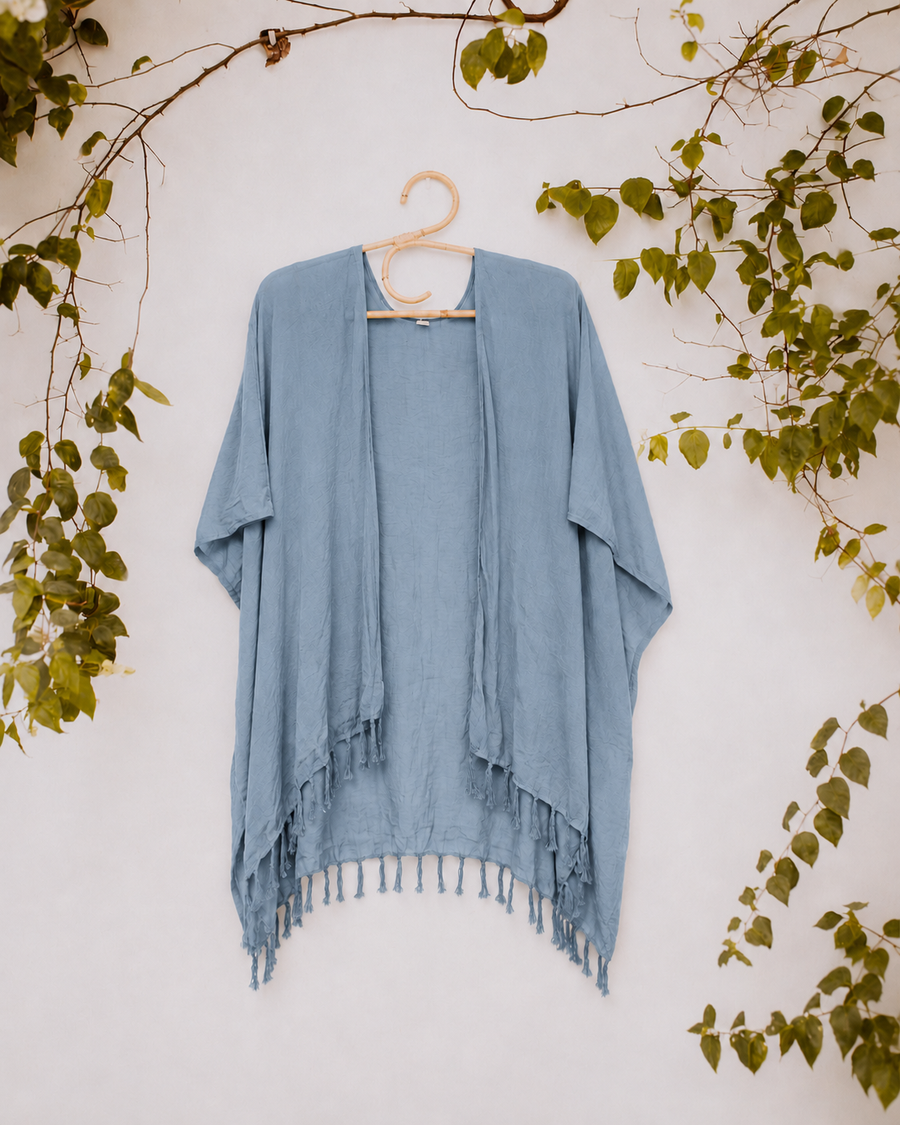 Soft slate blue Village Thrive Rayon Kimono with fringe hem on a hanger against a vine-covered white background.