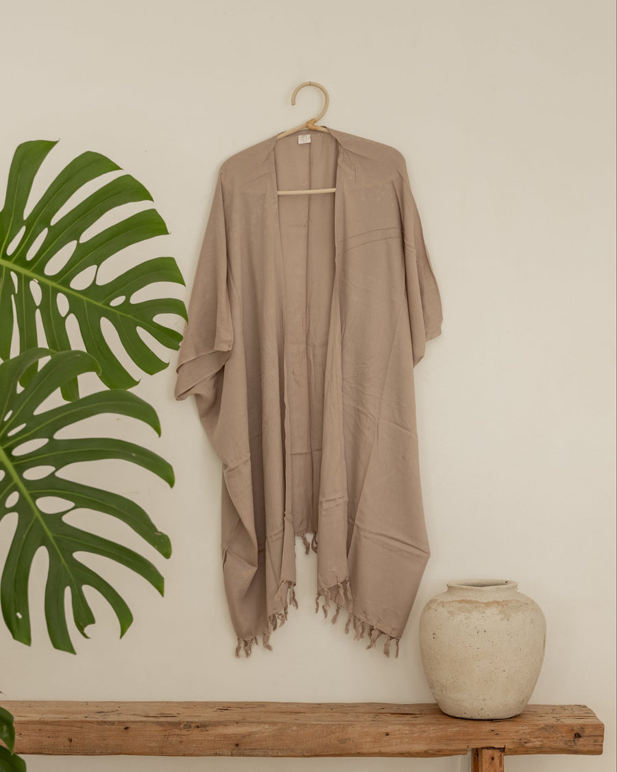 Taupe Village Thrive Rayon Kimono hanging on a bamboo hanger next to a Monstera plant and rustic pot.