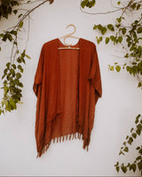 Terracotta Village Thrive Rayon Kimono with fringe trim hanging on a rattan hanger against a white wall with green vines.