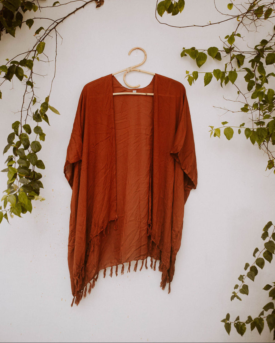 Terracotta Village Thrive Rayon Kimono with fringe trim hanging on a rattan hanger against a white wall with green vines.