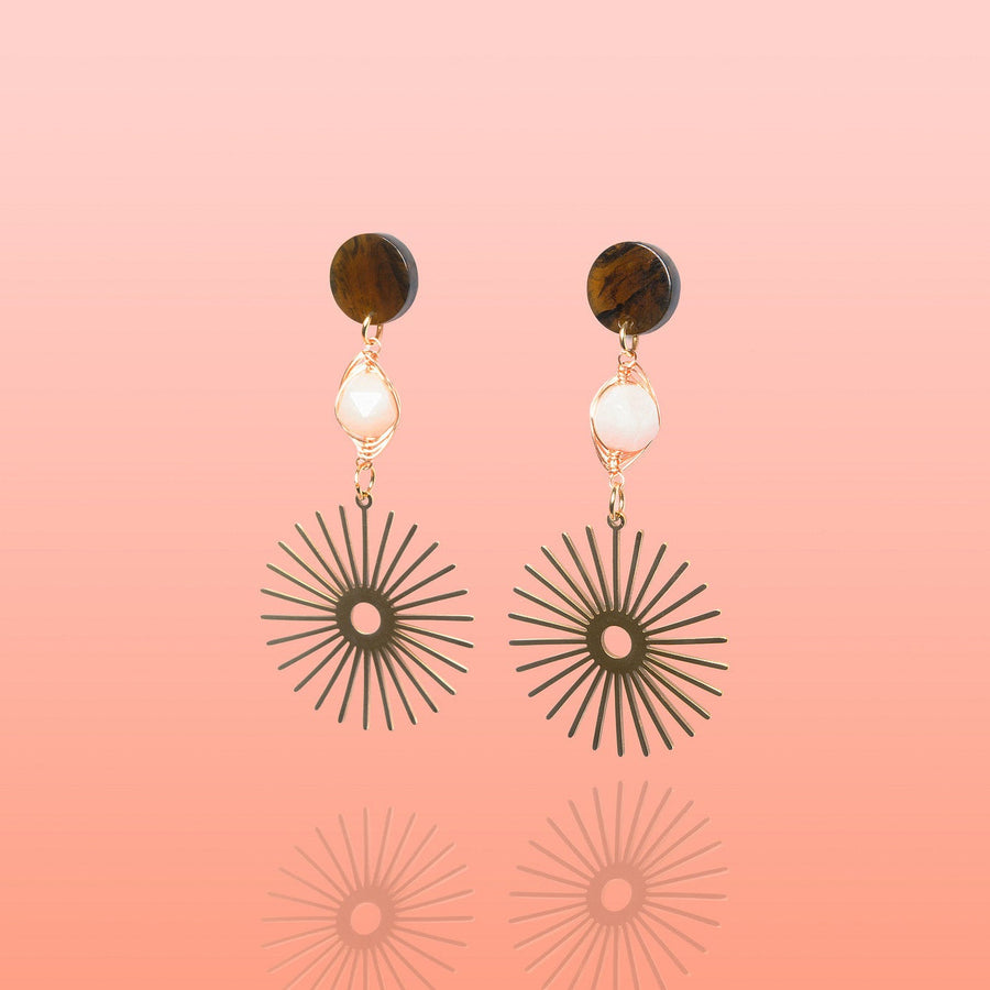 A pair of multi-tier Tribe and Temple Isela statement earrings displaying round marbled brown resin posts, wire-wrapped pink jade gemstones, and large radiant gold starburst charms against a soft coral pink gradient background.