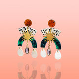 A pair of multi-tier Tribe and Temple Maribel Shell Statement Earrings showing gold sunburst connectors, green marbled resin arcs, and iridescent white shell drops against a soft coral pink gradient background.