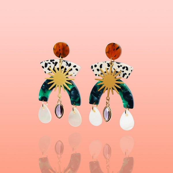 A pair of multi-tier Tribe and Temple Maribel Shell Statement Earrings showing gold sunburst connectors, green marbled resin arcs, and iridescent white shell drops against a soft coral pink gradient background.